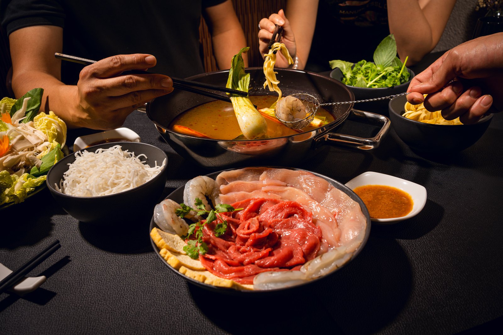 Vietnamesischer Hot Pot am Tisch: Br&uuml;he im Topf, Teller mit Rind, H&uuml;hnchen und Garnelen, Reisnudeln und frische Kr&auml;uter, G&auml;ste greifen mit St&auml;bchen zu